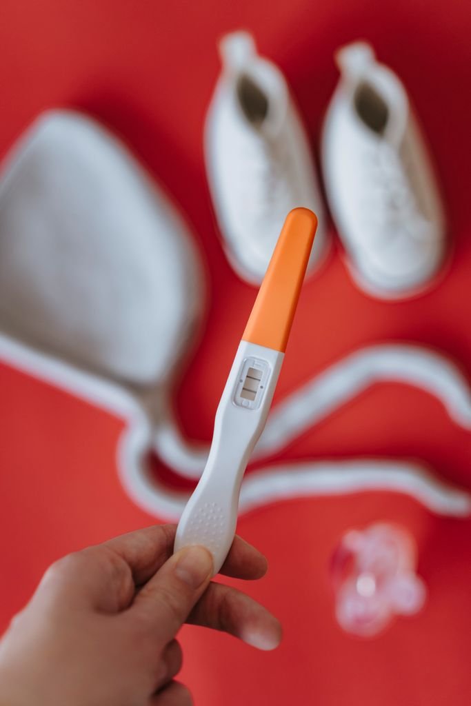 A positive pregnancy test holder alongside white baby shoes on a vibrant red surface.