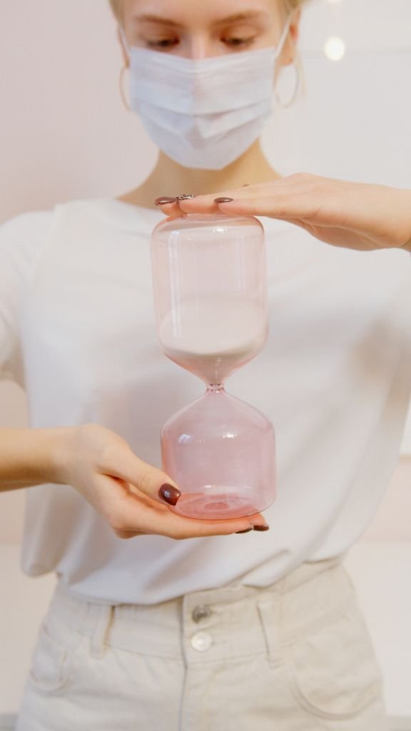 A woman wearing a facemask holds a pink hourglass, symbolizing time and health awareness.